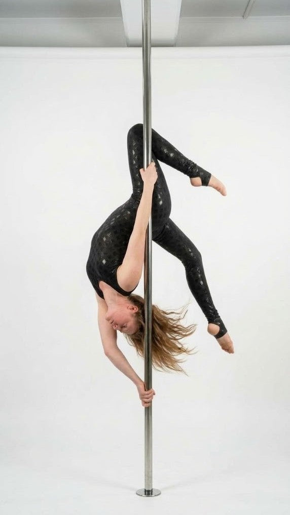 A woman in a black speckled pole dance leotard hangs upside down from a silver metal pole in a studio with a white background.
