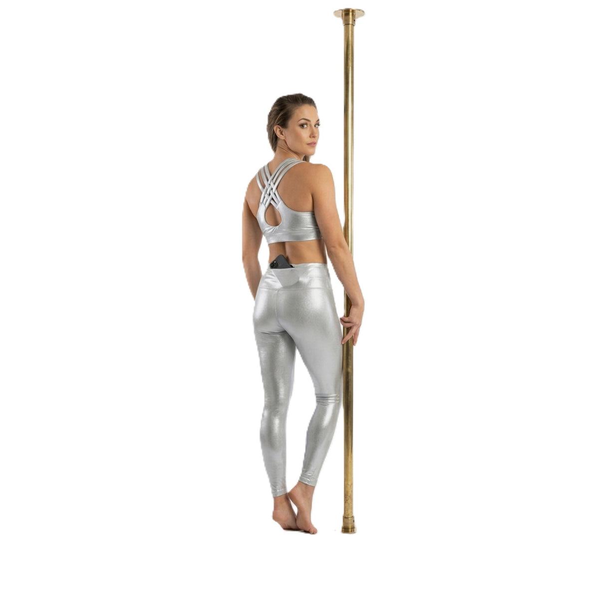 A woman stands barefoot against a vertical gold pole, wearing a shiny metallic silver sports bra and matching high-waisted leggings, shown from the back on a plain white background.