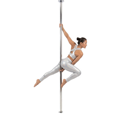 A woman in a silver strappy sports bra and matching shiny leggings poses on a vertical silver pole against a plain white background.